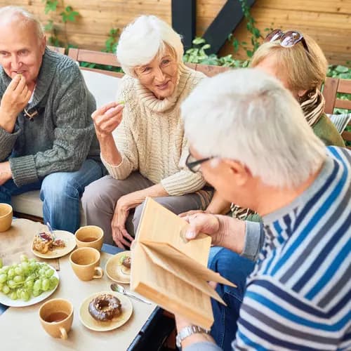Group of seniors sitting in outdoor patio enjoying coffee cake and fruit while reading a book