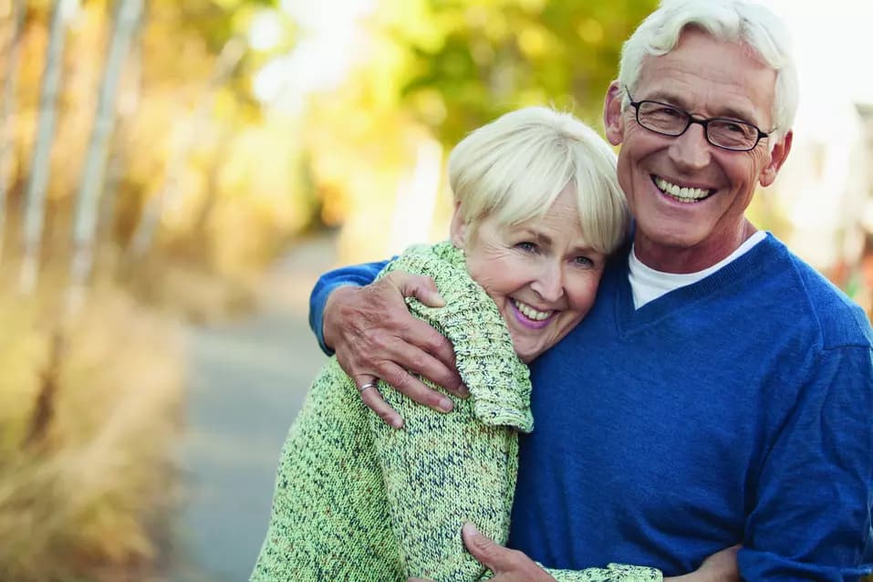 A senior couple hugging each other with big smile at autumn park trail / Un couple de personnes âgées s'étreignant avec un grand sourire sur un sentier dans un parc en automne