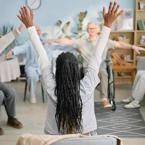 Instructor leading group exercise with senior participants seated in living room setting. / Un instructeur dirige un exercice collectif avec des participants âgés assis dans un salon.