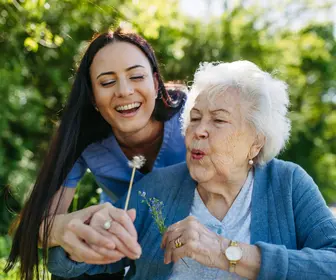 Caregiver and senior woman in wheelchair holding dandelion, picking wild flowers. Nurse and elderly woman enjoying a warm day in nursing home, public park.