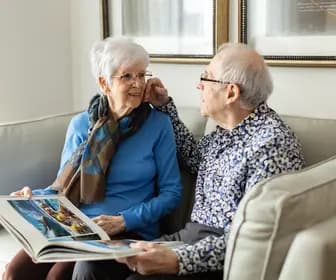 Joyful senior couple sitting on couch with a book/reading while looking at each other / Un couple de personnes âgées rayonnant de joie, assis sur un canapé, un livr