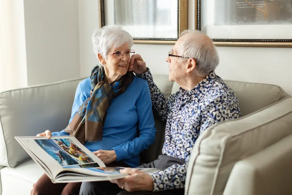 Joyful senior couple sitting on couch with a book/reading while looking at each other / Un couple de personnes âgées rayonnant de joie, assis sur un canapé, un livr