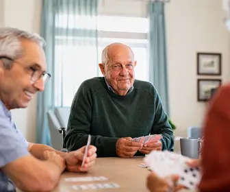 Retired senior man in wheelchair playing cards with friends at nursing home. Smiling group of old people playing cards at care centre. Happy and content seniors playing at lunch table and havin fun together.