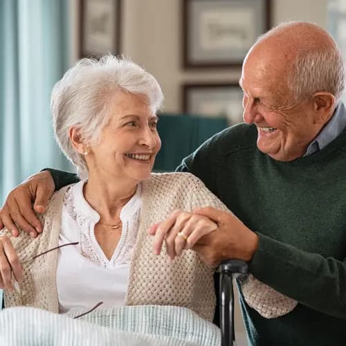 Romantic senior husband embracing disabled wife sitting on wheelchair at nursing home. Cheerful old couple embracing and looking at each other with love. Happy elderly man holding hands of handicapped woman sitting in wheelchair at care centre.