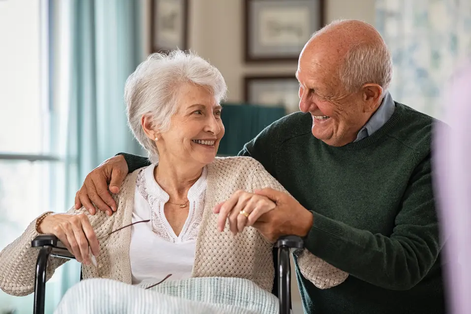 Romantic senior husband embracing disabled wife sitting on wheelchair at nursing home. Cheerful old couple embracing and looking at each other with love. Happy elderly man holding hands of handicapped woman sitting in wheelchair at care centre.