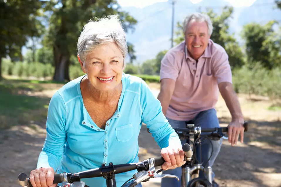 Senior couple rides bike on a gravel road at park under sun / Un couple de personnes âgées fait du vélo sur un chemin de gravier dans un parc, sous le soleil