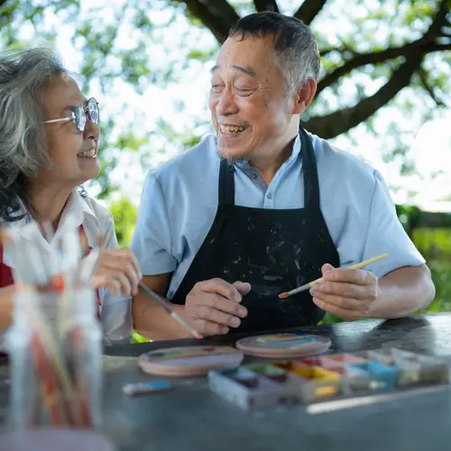 Senior asian couple painting pottery together at a table outside.