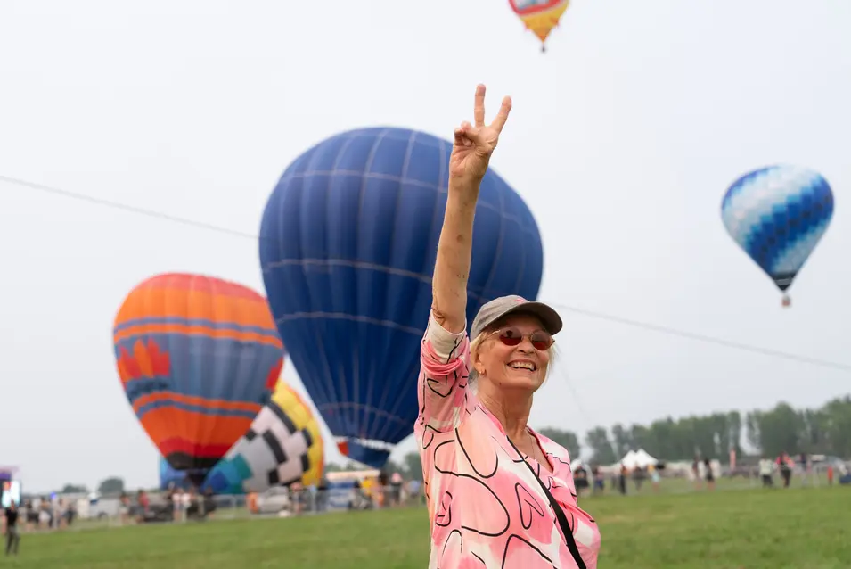 Senior woman who got her wishes granted through Chartwell Wish of a Lifetime poses V hand gesture in front of air balloons / Une aînée dont les vœux ont été exaucés grâce à Chartwell Wish of a Lifetime fait un geste de la main devant des ballons gonflables