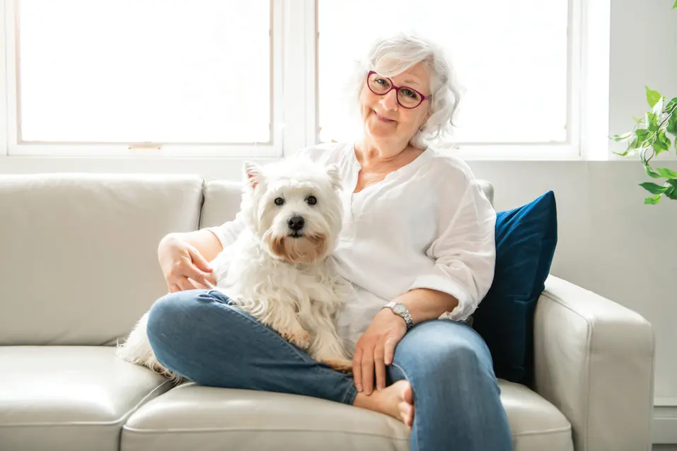 Senior woman is sitting on her couch along with her white dog / Une femme âgée est assise sur son canapé avec son chien blanc.