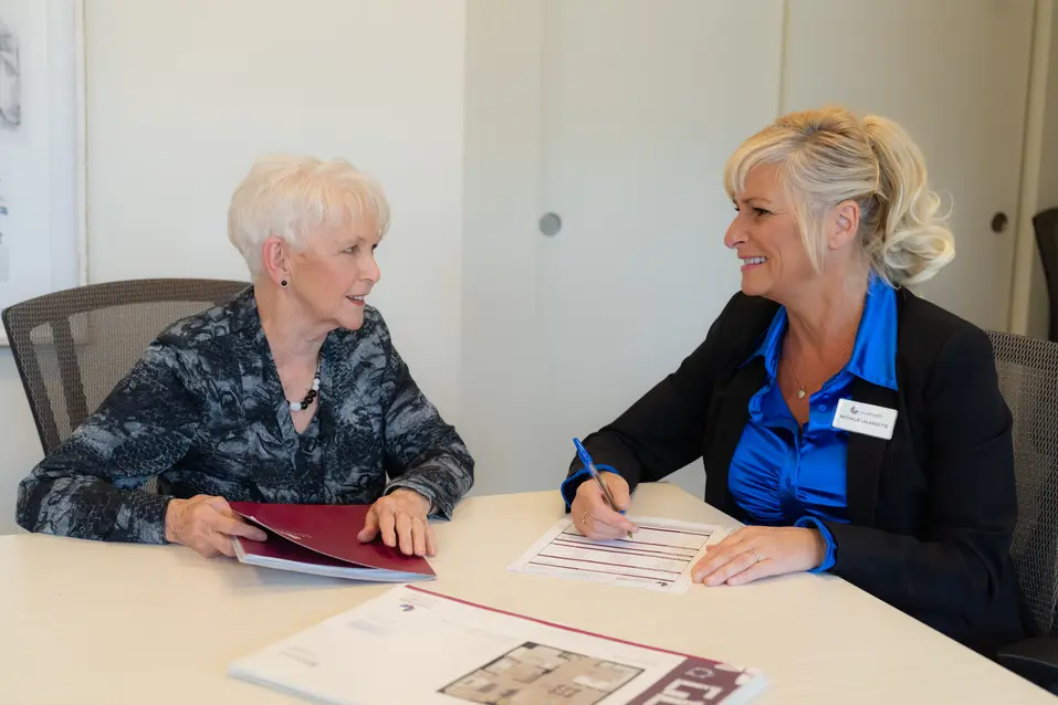 Senior woman sits down with Chartwell staff to do a consultation. Staff is sitting down writing on a piece of paper / Une femme âgée s'assoit avec le personnel de Chartwell pour une consultation. Le personnel est assis et écrit sur une feuille de papier