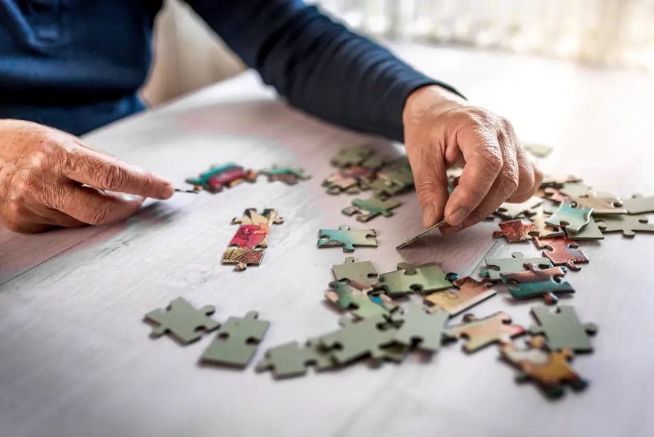 Closeup of the old hands playing with jigsaw puzzles at the white table at home. Connecting pieces together.