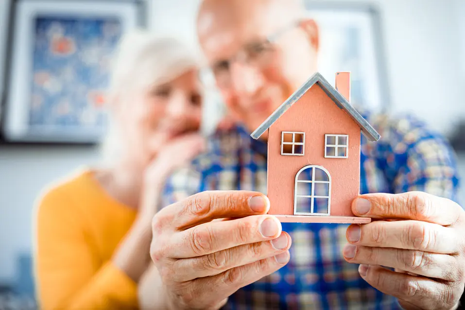 Senior couple is showing up a pink coloured wooden house model / Un couple âgé montre une maquette de maison en bois de couleur rose