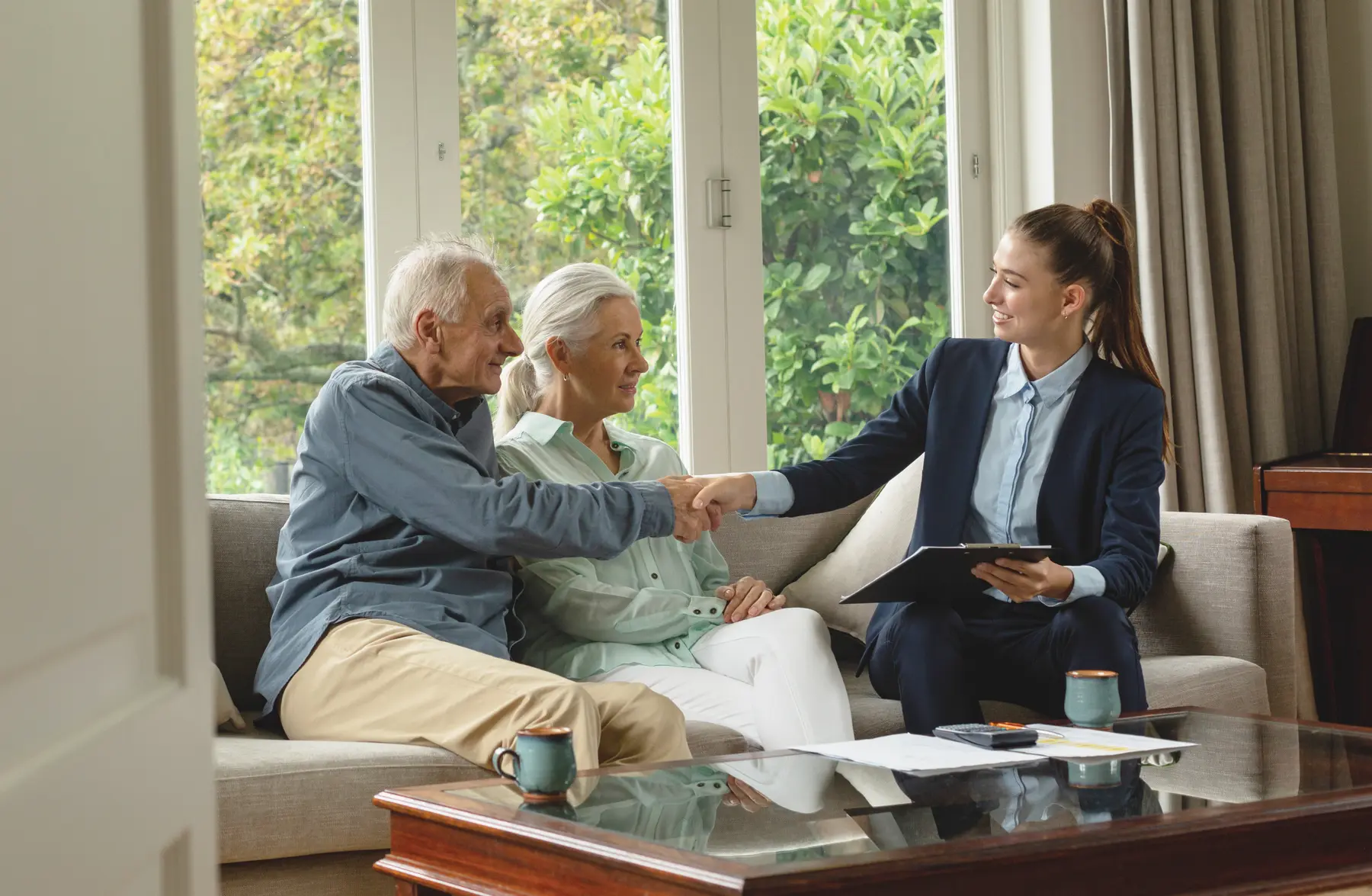 Senior couple sitting in couch is shaking hand of a female real estate agent in suit / Un couple de seniors assis dans un canapé serre la main d'un agent immobilier en tailleur