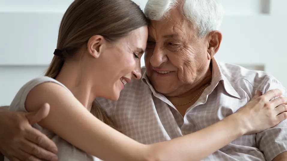 Loving adult daughter and older father are hugging and touching foreheads with gentle smiles / Une fille adulte aimante et un père plus âgé se serrent dans les bras et se touchent le front en souriant doucement