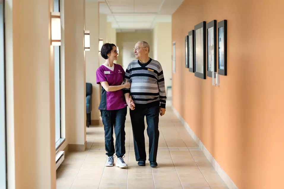 Chartwell staff is assisting senior male resident walking down an aisle / Le personnel de Chartwell aide un résident âgé à marcher dans l'allée