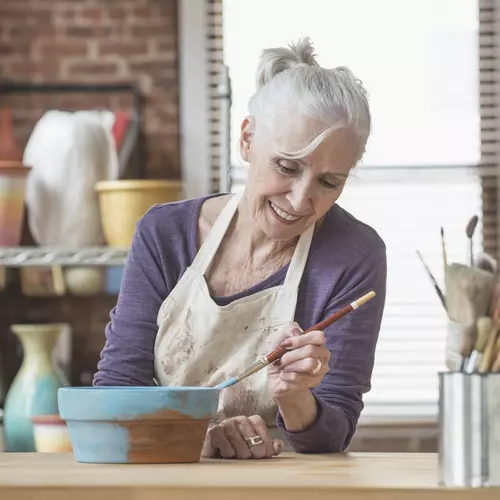 Senior woman in a studio painting a pot of cloy with a brush
