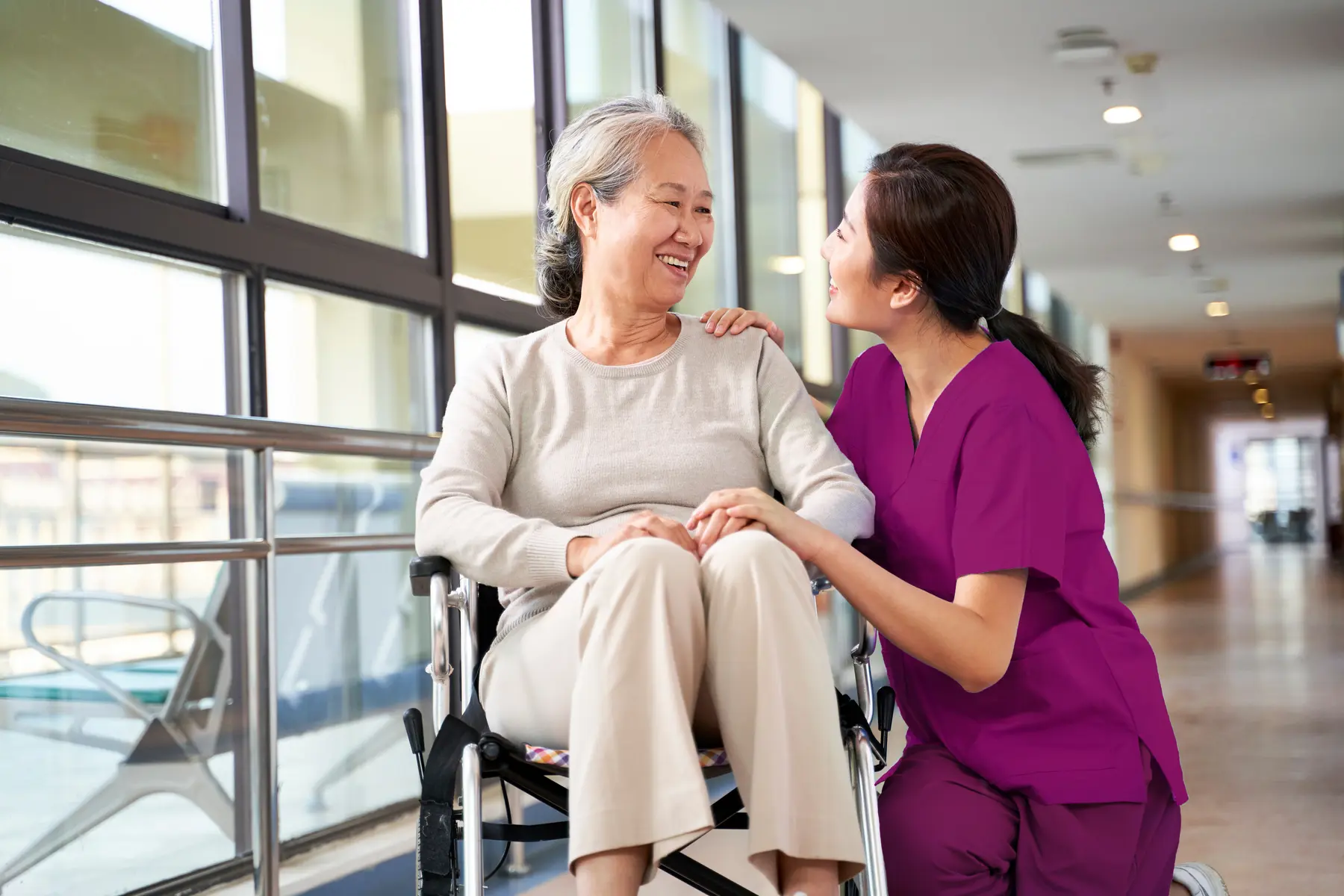 Senior woman in wheelchair and care giver are having conversation with smile / Une femme âgée en fauteuil roulant et une aide-soignante discutent en souriant
