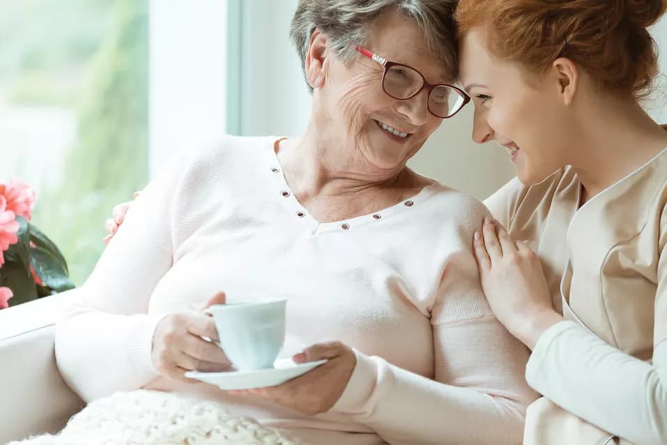 Elder woman with cup of tea and her friendly caretaker looking in each other's eyes / Femme âgée avec une tasse de thé et sa gardienne amicale se regardant dans les yeux